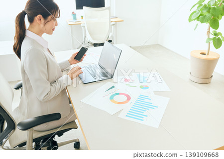 A young female business person checking documents using her smartphone and laptop in the office. 139109668