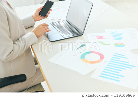 A businesswoman analyzing graph data using her smartphone and laptop in the office. 139109669
