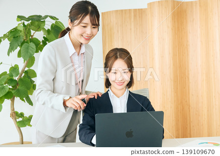 A young female employee receiving guidance from a senior colleague while looking at a laptop in the office - On-the-Job Training (OJT) scene. 139109670