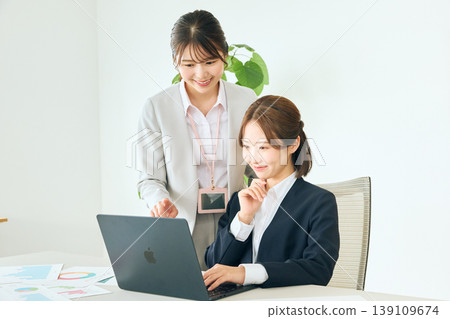 兩位年輕的女性商務人士正在辦公室開會，她們各自看著筆記型電腦——一位是資深同事，另一位是初級同事。 139109674