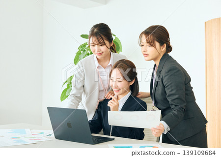 三位年輕的女性商務人士正在辦公室開會，她們看著筆記型電腦和文件，展現出團隊合作。 139109684