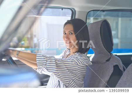 A Japanese woman is sitting in the driver's seat of a car, holding the steering wheel and smiling at the camera. A Japanese woman is sitting in the driver's seat of a car, holding the steering wheel and smiling at the camera. 139109939