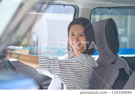 A Japanese woman is sitting in the driver's seat of a car, holding the steering wheel and smiling at the camera. 139109940
