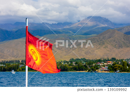 National flag of Kyrgyzstan on pole by serene Issyk-Kul lake with mountains and village National flag of Kyrgyzstan on pole by serene Issyk-Kul lake with mountains and village 139109992