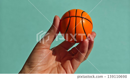 Hand holds a small orange basketball against a light blue background in a simple setting 139110698
