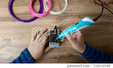 Child is using a 3D pen to create a small house model on a wooden surface with colorful wires Child is using a 3D pen to create a small house model on a wooden surface with colorful wires 139110700