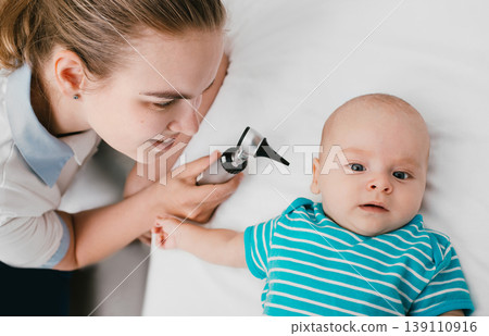 Smiling pediatrician doing ear exam with an otoscope to a baby at the hospital. Doctor examines infant baby 3-month-old Smiling pediatrician doing ear exam with an otoscope to a baby at the hospital. Doctor examines infant baby 3-month-old 139110916