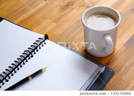 The image is of someone working at their desk, with a notebook open, while sipping a latte (caffè latte) with plenty of milk. The image is of someone working at their desk, with a notebook open, while sipping a latte (caffè latte) with plenty of milk. 139110988