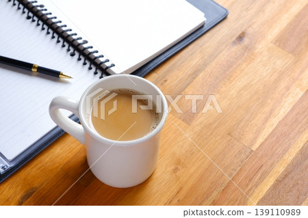 The image is of someone working at their desk, with a notebook open, while sipping a latte (caffè latte) with plenty of milk. The image is of someone working at their desk, with a notebook open, while sipping a latte (caffè latte) with plenty of milk. 139110989