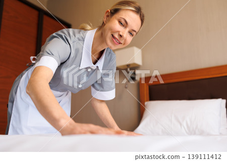 Close up of smiling hotel housekeeper making neatly arranged bed Close up of smiling hotel housekeeper making neatly arranged bed 139111412
