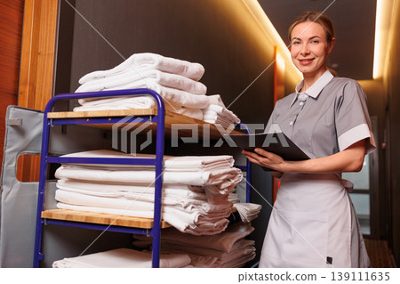 Smiling Housekeeper Holding Clipboard Next to Linen Cart in Hotel Corridor Smiling Housekeeper Holding Clipboard Next to Linen Cart in Hotel Corridor 139111635