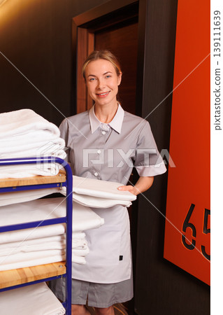 Friendly housekeeping staff member with linen cart standing in luxury hotel hallway near guest room 139111639