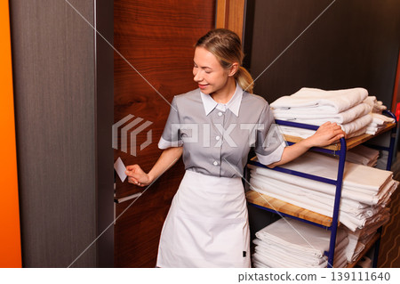 Hotel housekeeper opening guest room door with key card while standing beside fully loaded linen cart 139111640
