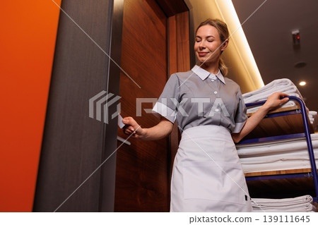 Low angle portrait of hotel maid opening room door beside housekeeping cart with clean laundry 139111645