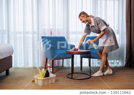 Hotel maid disinfecting wooden table with spray bottle between blue armchairs Hotel maid disinfecting wooden table with spray bottle between blue armchairs 139111662