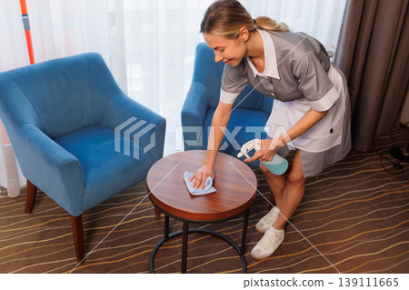 Top view maid sanitizing wooden coffee table beside blue armchair in hotel room Top view maid sanitizing wooden coffee table beside blue armchair in hotel room 139111665