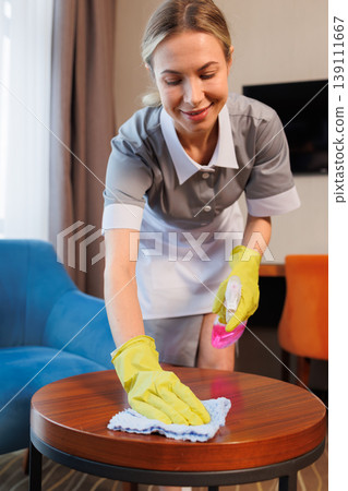 Smiling female housekeeper wearing gloves cleaning wooden table with spray bottle in hotel room Smiling female housekeeper wearing gloves cleaning wooden table with spray bottle in hotel room 139111667