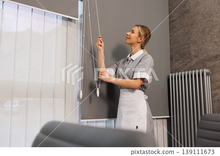 Smiling housekeeper standing near roller blinds in corporate meeting room with chairs 139111673