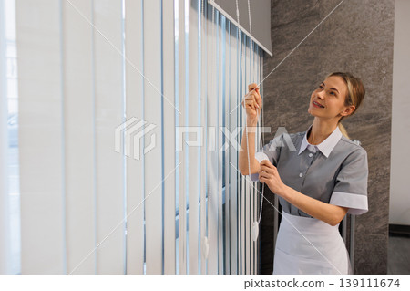 Female cleaning staff opening roller blinds in modern office interior with radiator and window light 139111674