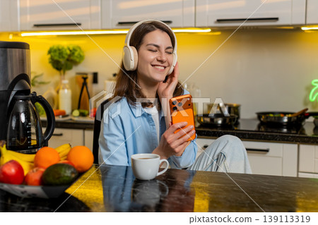 Young woman listening to music with headphones using smartphone in home kitchen relaxing with coffee Young woman listening to music with headphones using smartphone in home kitchen relaxing with coffee 139113319