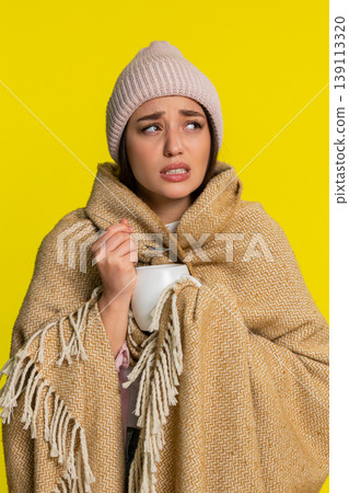 Woman wrapped in blanket wearing hat adding honey to tea slowly drinking caring for sore throat 139113320