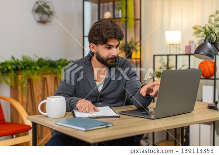 Indian man cross checks numbers on laptop versus paper reports focused data review serious look calm Indian man cross checks numbers on laptop versus paper reports focused data review serious look calm 139113353