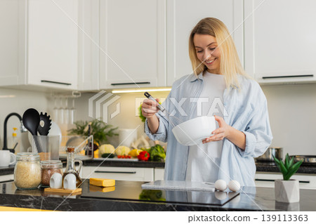 Young woman whisking eggs in bowl in modern home kitchen preparing omelet batter for quick breakfast 139113363