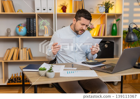 Young man on home table checking bills and reacting to unexpected payments with frustration stress Young man on home table checking bills and reacting to unexpected payments with frustration stress 139113367