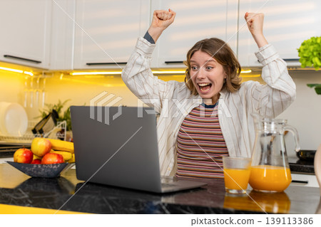 Young woman celebrating online success with laptop in home kitchen during breakfast with raised arms Young woman celebrating online success with laptop in home kitchen during breakfast with raised arms 139113386