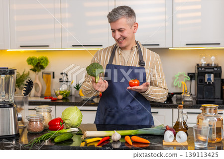 Middle-aged man comparing broccoli and tomato in kitchen while choosing vegetables for healthy meal 139113425