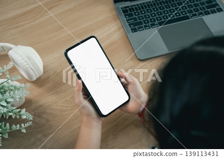 Person holding smartphone with blank screen above wooden table featuring laptop, headphones, and plant on background in modern workspace setting 139113431