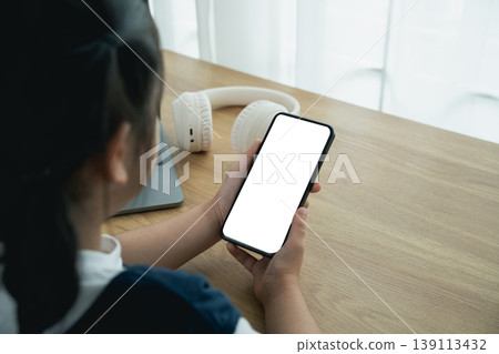Young girl holding smartphone with blank screen, headphones on table, working on a wooden desk in a bright indoor space with soft natural light Young girl holding smartphone with blank screen, headphones on table, working on a wooden desk in a bright indoor space with soft natural light 139113432