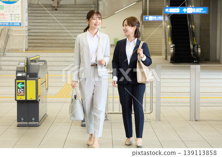 兩位身著職業套裝的年輕女商人走過火車站的檢票口；這是上班族通勤途中面帶微笑的場景。 139113858