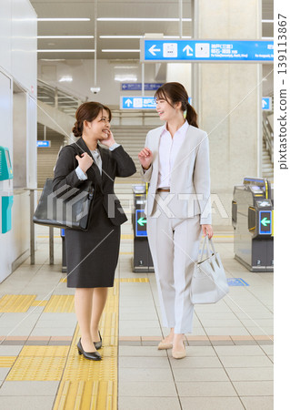 Two young women in suits, business professionals, talking near the ticket gates of a train station - a commuting image. 139113867