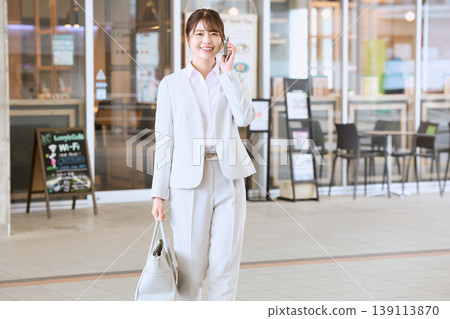 一位身穿白色套裝的年輕女商務人士邊走邊用智慧型手機通話；這是銷售人員在路上的畫面。 139113870