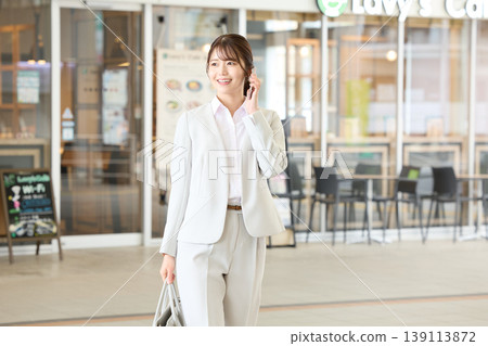 一位身穿白色職業套裝的年輕日本女商人一邊用智慧型手機通話,一邊在辦公區的一家咖啡館前行走。 一位身穿白色職業套裝的年輕日本女商人一邊用智慧型手機通話,一邊在辦公區的一家咖啡館前行走。 139113872