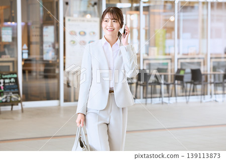 A young woman in a white suit, a business person, walks while talking on her smartphone, smiling in front of a cafe in the office district. 139113873