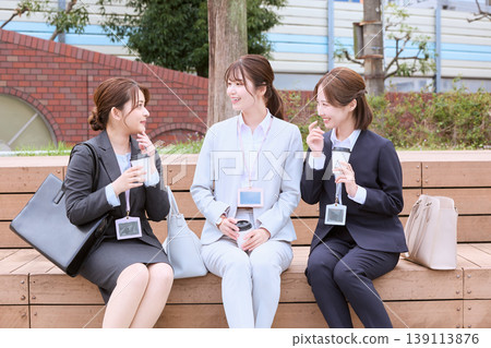 三位年輕的職業婦女在戶外長椅上一邊喝咖啡一邊聊天;她們是正在午休的上班族。 三位年輕的職業婦女在戶外長椅上一邊喝咖啡一邊聊天;她們是正在午休的上班族。 139113876