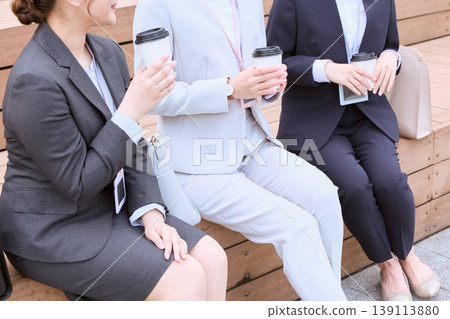 Three businesswomen in suits are chatting on an outdoor bench, holding coffee cups. Three businesswomen in suits are chatting on an outdoor bench, holding coffee cups. 139113880