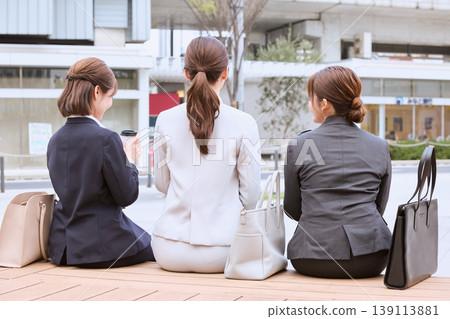 Three businesswomen in suits sitting on a bench in the city; seen from behind; office workers on break; job hunting. Three businesswomen in suits sitting on a bench in the city; seen from behind; office workers on break; job hunting. 139113881