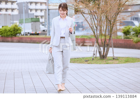 一位身穿淺灰色褲裝的年輕女商人，在春日的城市街道上輕快地行走，給人一種銷售或通勤的印象。 139113884