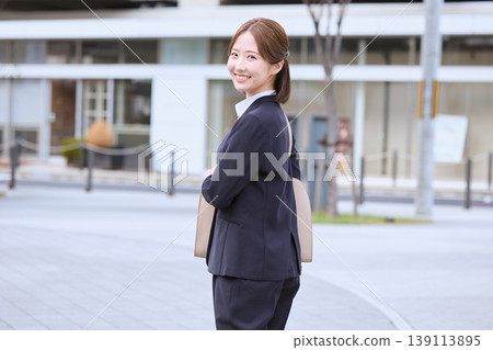 A young female businessperson in a suit, looking back and smiling; a new employee walking through an office district; an image of job hunting. A young female businessperson in a suit, looking back and smiling; a new employee walking through an office district; an image of job hunting. 139113895