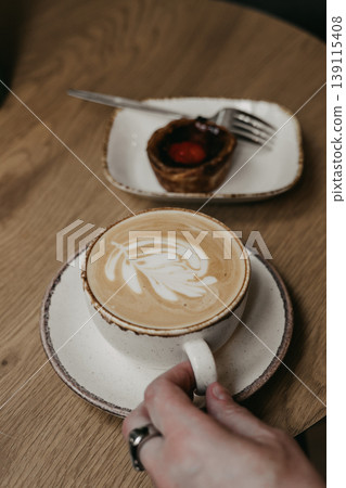 Close up of person hand holding cup of cappuccino with latte art and pastel de nata Portugal dessert on wooden table. Cafe morning, breakfast 139115408