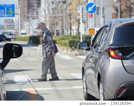 A scene of cars waiting at a traffic light on a city road during a commute, and a senior citizen crossing a pedestrian crossing. A scene of cars waiting at a traffic light on a city road during a commute, and a senior citizen crossing a pedestrian crossing. 139116704