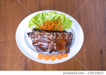 Overhead view of grilled BBQ pork ribs on a white plate, glazed with savory sauce. Garnished with fresh lettuce and carrots on a rustic wooden background. Delicious meat dish for food photography. Overhead view of grilled BBQ pork ribs on a white plate, glazed with savory sauce. Garnished with fresh lettuce and carrots on a rustic wooden background. Delicious meat dish for food photography. 139119027