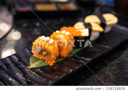 Assorted sushi platter featuring California rolls with orange fish roe and egg tamago nigiri on a black plate. Fresh Japanese cuisine served in a restaurant setting with elegant bokeh lighting. 139119029