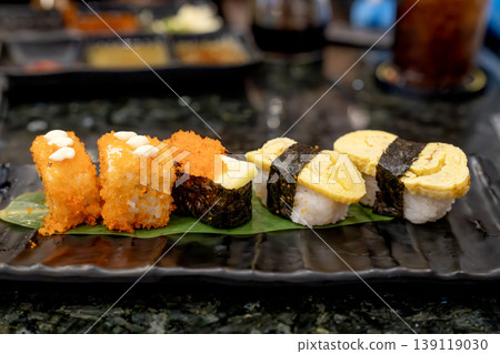 Assorted sushi platter featuring tamago nigiri, tobiko gunkan maki, and roe-covered rolls on a black plate in a Japanese restaurant. Close-up view of fresh seafood appetizers on dark table. Assorted sushi platter featuring tamago nigiri, tobiko gunkan maki, and roe-covered rolls on a black plate in a Japanese restaurant. Close-up view of fresh seafood appetizers on dark table. 139119030
