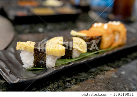 Assorted Japanese sushi platter featuring tamago nigiri with egg omelet and nori seaweed, followed by rolls with fish roe. Close-up shot on black plate in restaurant setting. Delicious Asian cuisine. Assorted Japanese sushi platter featuring tamago nigiri with egg omelet and nori seaweed, followed by rolls with fish roe. Close-up shot on black plate in restaurant setting. Delicious Asian cuisine. 139119031