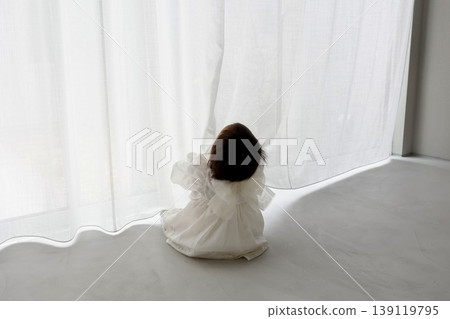 The back view of a one-year-old child in a white dress and white curtains. A toddler gazing out the window in the light. 139119795