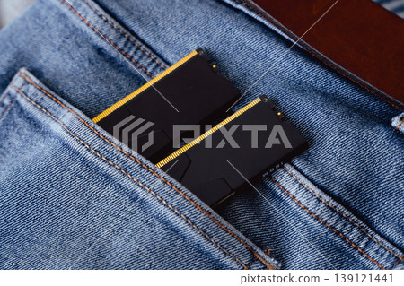 Black DDR5 RAM memory modules placed inside a jeans pocket. RAM shortages on the market, rising prices, and increased demand driven by the growing need for AI training. Black DDR5 RAM memory modules placed inside a jeans pocket. RAM shortages on the market, rising prices, and increased demand driven by the growing need for AI training. 139121441